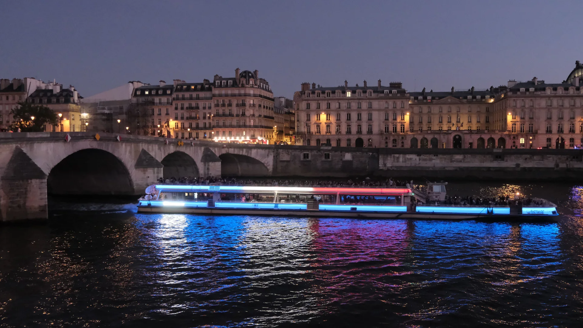 A Bateaux Mouches boat lit in blue, white and red, cruising the Seine at dusk past a stone bridge and the lit Left Bank quays
